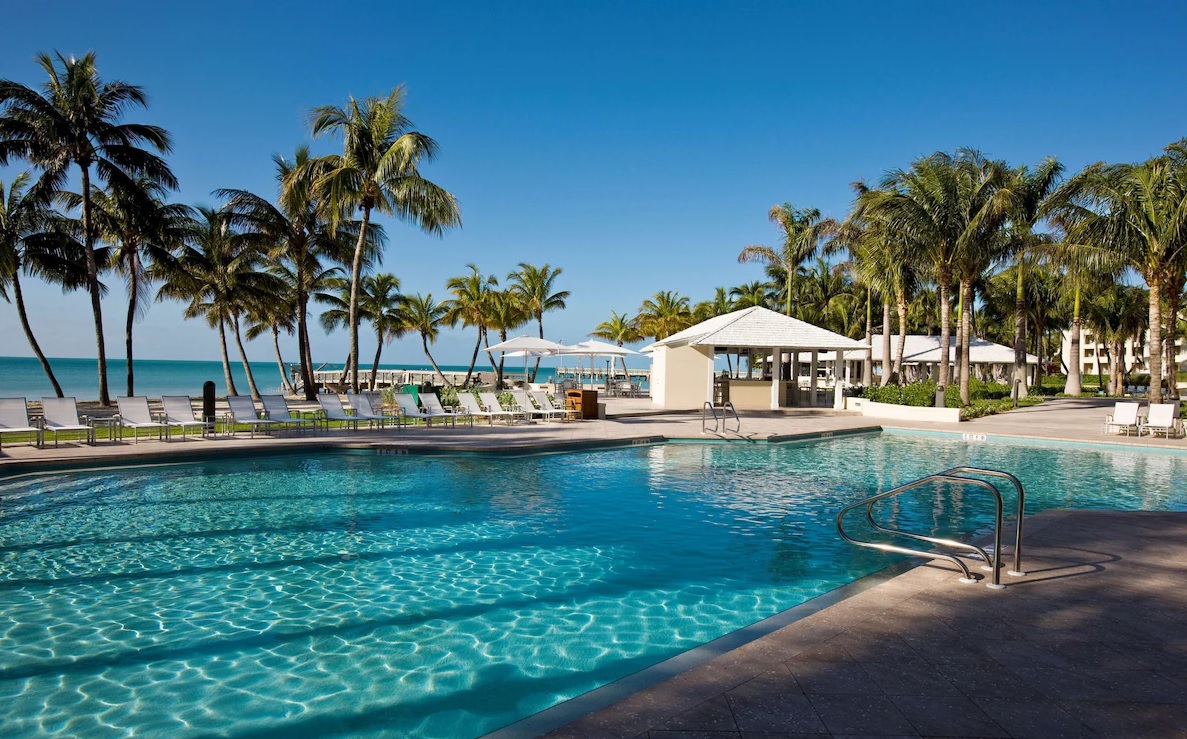 A serene tropical resort pool area features clear blue water, surrounded by tall palm trees and lounge chairs. An ocean is visible in the background under a clear blue sky, with a few white cabanas providing shade near the sandy beach. This luxurious experience is reminiscent of Casa Marina Key West.