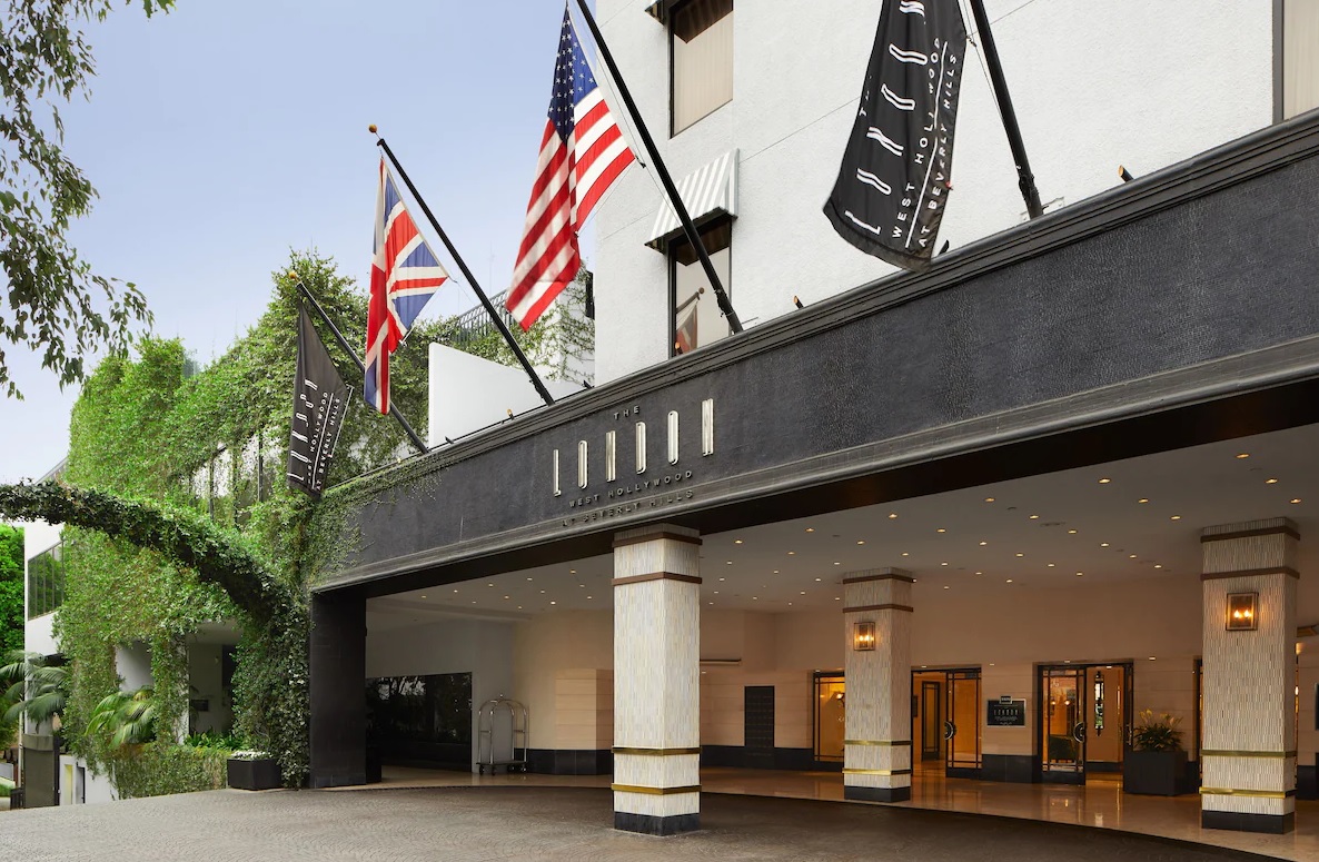 Entrance of a hotel adorned with the UK's Union Jack and the US flag, each positioned prominently on either side. The hotel has a sophisticated entrance canopy with the name "London West Hollywood" visible. Black flags with white text also hang beside the national flags, exuding luxury.