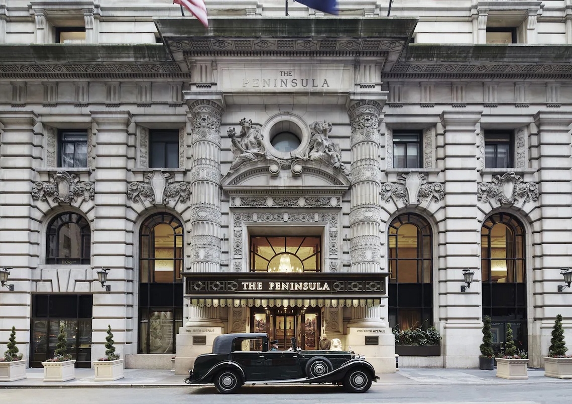 A grand building with intricate stone detailing, arched windows, and a large entrance covered by a marquee labeled "The Peninsula New York." A vintage black car is parked in front, and there are flags flying above the entrance. The hotel promises luxury with its acclaimed Rooftop Experience.