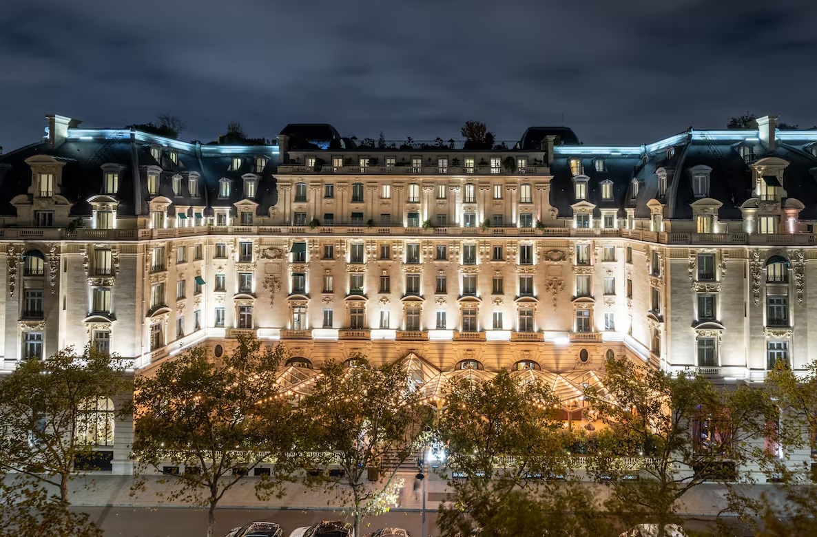 A grand, multi-story hotel building illuminated at night exudes luxurious elegance. The ornate façade of The Peninsula Paris features numerous windows, balconies, and arches, all lit warmly. Trees with autumn foliage line the street in front of this 16th Arrondissement gem, with parked cars visible at the bottom of the image.