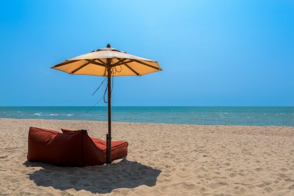 A peaceful beach scene featuring a sandy shore, calm blue ocean, and a clear sky. A brown lounge chair and a large beige umbrella sit on the sand, offering a shaded spot to relax and enjoy the surroundings—truly a dream getaway that rivals top Thailand hotels.