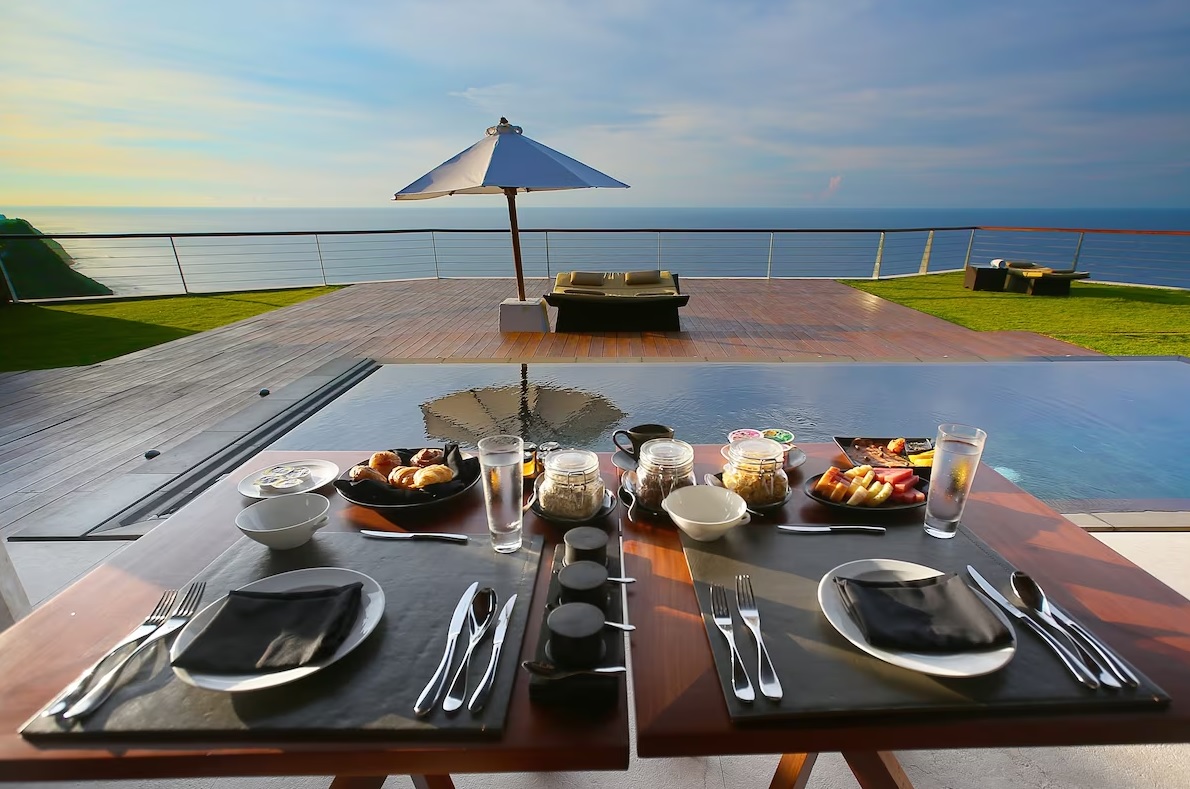 A beautifully set table for two overlooks a vast ocean view. The table is set with black placemats, cutlery, plates, and glasses of water, accompanied by a variety of food items. In the background, an umbrella shades a sun lounger on a wooden deck with green grass. The Edge Bali truly epitomizes luxury accommodation.