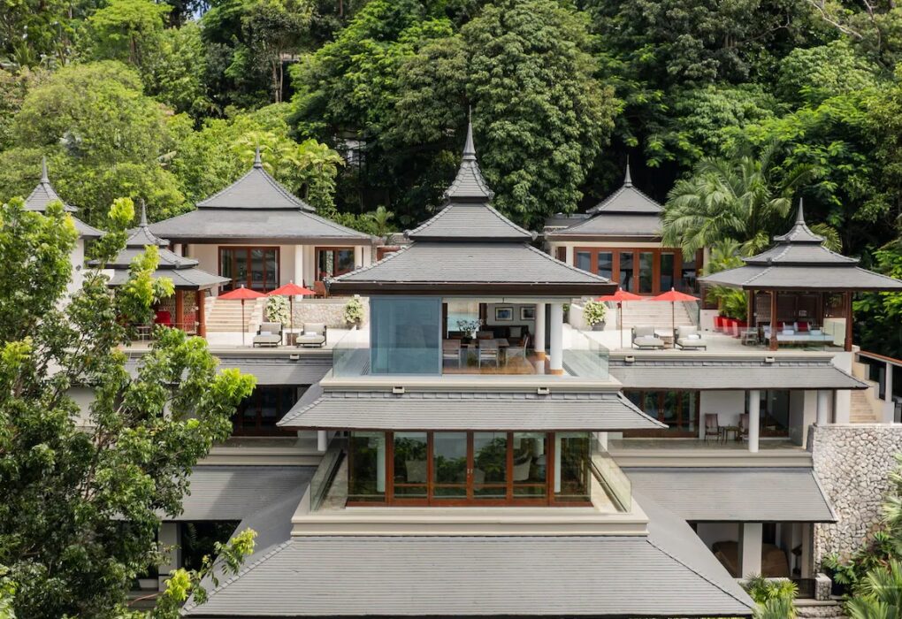 A luxurious multi-level villa surrounded by lush greenery, Trisara Villas in Phuket features traditional Asian architectural elements, such as tiered roofs and wooden accents. Spacious terraces and balconies offer outdoor seating with multiple lounge areas and red umbrellas, perfect for a luxury escape.