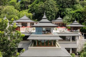 A luxurious multi-level villa surrounded by lush greenery, Trisara Villas in Phuket features traditional Asian architectural elements, such as tiered roofs and wooden accents. Spacious terraces and balconies offer outdoor seating with multiple lounge areas and red umbrellas, perfect for a luxury escape.